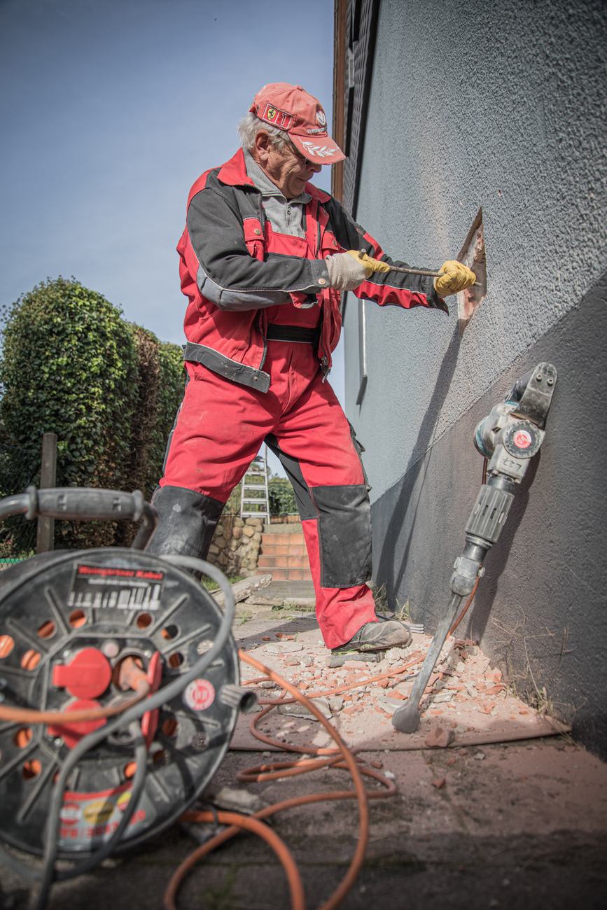 Handwerker in roter Arbeitskleidung öffnet mit einem Meißel die Fassade, daneben ein Abbruchhammer