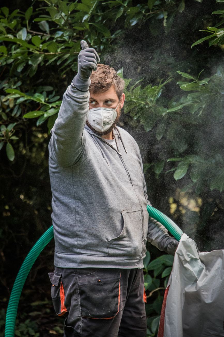 Mitarbeiter mit Atemschutzmaske zeigt den Daumen nach oben, daneben ein grüner Einblasschlauch