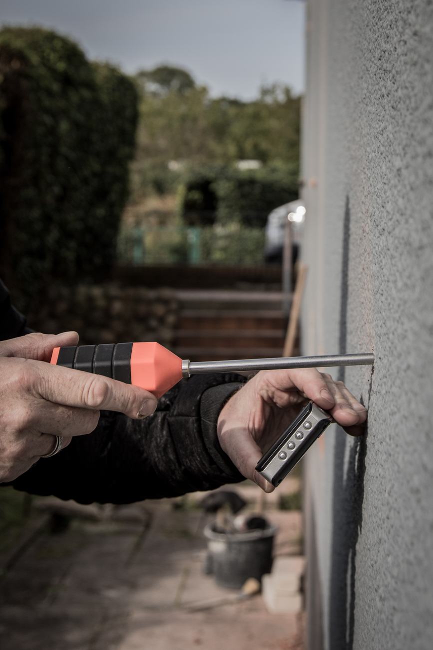 Zwei Hände halten eine Sonde und ein Messgerät an eine verputzte Hauswand